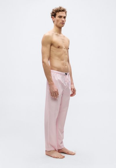 Νέος άνδρας χωρίς μπλούζα, όρθιος, ξυπόλητος, φορώντας άνετο ροζ και λευκό ριγέ παντελόνι πυτζάμας, μπροστά σε απλό ανοιχτόχρωμο φόντο.