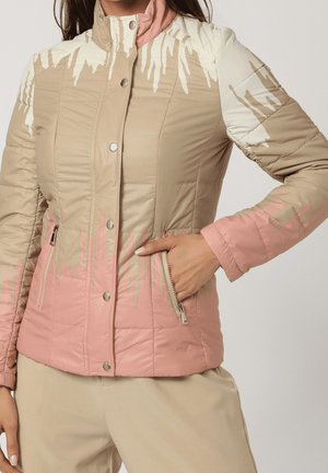 Mujer con una chaqueta acolchada beige y rosa con diseño de goteo de pintura blanca, mano en el bolsillo con cremallera, combinada con pantalones beige.