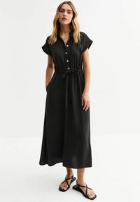 Vestido midi negro con mangas cortas, botones en la parte delantera, cintura ajustable con cordón y bolsillos laterales. Tejido suave con corte relajado y estilo hasta el tobillo.