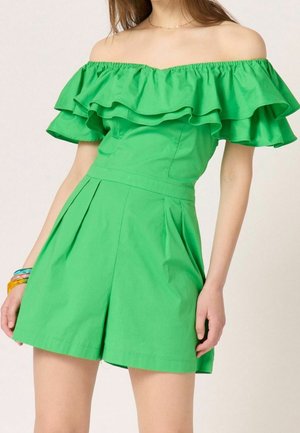 Kvinde iført en klar grøn off-shoulder kjole med flæseærmer og plisserede shorts, accessoriseret med farverige armbånd på det ene håndled.