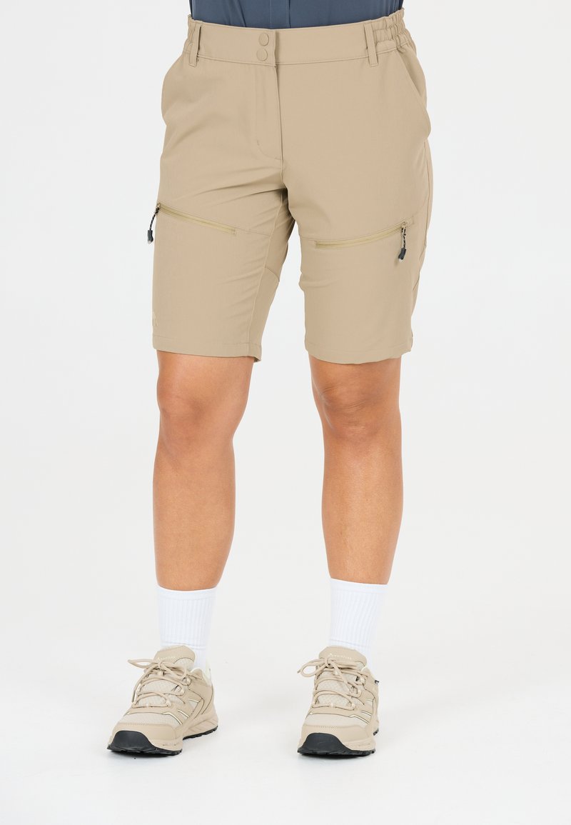 Personne portant un short beige arrivant aux genoux avec des poches zippées sur les cuisses, des baskets beiges et des chaussettes blanches montantes sur un fond blanc uni.