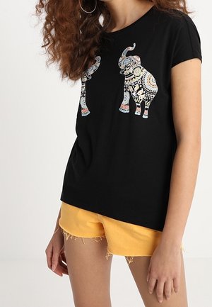 Femme portant un t-shirt noir avec des motifs colorés d'éléphants et un short jaune effiloché, debout devant un fond clair uni.