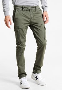 Olivenfarbene Cargo-Hosen aus leichtem Stoff mit mehreren Taschen und Nähdetails, kombiniert mit weißen Sneakers.