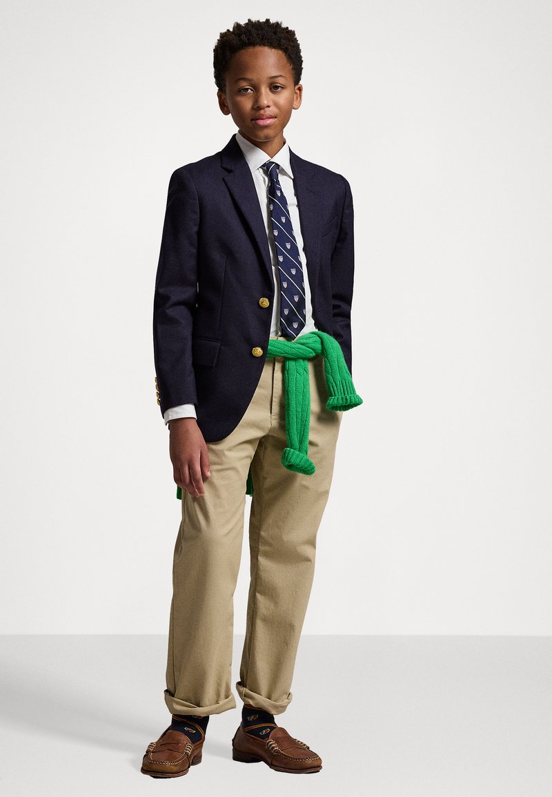 Blazer azul marino con botones dorados sobre una camisa blanca, combinado con pantalones caqui. Jersey verde atado a la cintura y mocasines marrones.