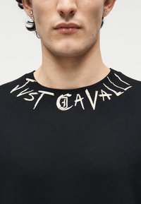 Νεαρό άτομο με μαύρο πουλόβερ με λαιμόκοψη, στο οποίο αναγράφεται η φθαρμένη λευκή επιγραφή «Just Cavalli» γύρω από τον γιακά, ορατό από τους ώμους μέχρι τα χείλη.