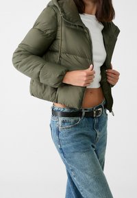 Olivgrön vadderad jacka med huva, dragkedja och resår i ärmslut, kombinerad med högmidjade blå denimjeans och en vit topp.