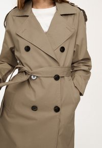 Trench-coat beige à double boutonnage, boutons noirs, ceinture à nouer, et épaulettes. Confectionné dans un tissu lisse et léger.