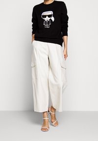 Sudadera negra con un personaje de dibujos animados con gafas de sol, combinada con pantalones cargo beige de pierna ancha y sandalias blancas con tiras.