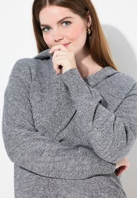 Grå stickad huvtröja i ett texturerat tyg, med en avslappnad passform och en vikbar krage. Synliga sömdetaljer och långa ärmar finns.