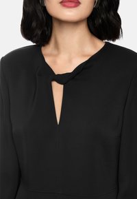 Schwarzes Kleid mit einem Schlüsselloch-Ausschnitt und Knoten-Detail, langen Ärmeln und einer glatten, weichen Textur. Minimalistisches und klassisches Design.