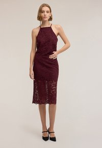 Vinröd spetsklänning med halterneck, figurnära design och midjelång fåll. Genomskinlig spetsstruktur med vågiga kanter. Svarta högklackade skor.