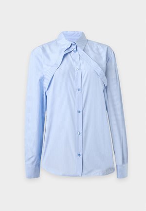 Femme portant une chemise ample bleu clair à rayures par-dessus un crop top blanc, un pantalon large beige et des sandales noires à talons, debout devant un fond uni.