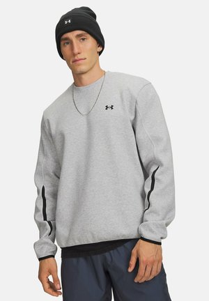 Young man wearing a gray Under Armour sweatshirt, black beanie, blue pants, and a silver chain necklace, standing against a white background.