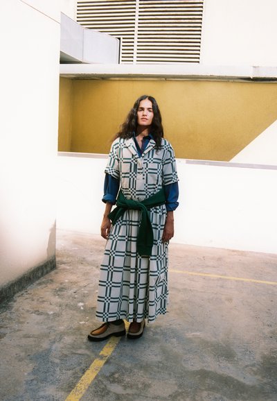 Une femme se tient dans une cour en béton, vêtue d’une longue robe à carreaux par-dessus une chemise bleue, avec un pull vert noué autour de la taille.