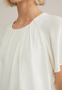 Blouse blanche texturée avec un col rond et des manches courtes, présentant de douces plis sur le devant. Tissu léger avec un éclat subtil.
