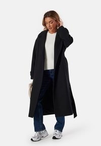 Cappotto lungo nero con taglio dritto e grandi revers, indossato sopra un maglione bianco e jeans blu. Il look è completato da sneakers bianche.