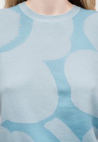 Ανοιχτό μπλε πλεκτό πουλόβερ με μεγάλες, ακανόνιστες στρογγυλεμένες σχηματισμούς σε λίγο πιο ανοιχτό μπλε χρώμα, που φοριέται από ένα άτομο.