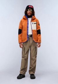 Oranža flīsa jaka ar melniem akcentiem un violetu apmali, bēšas cargo bikses un melni sporta apavi. Valkāts ar cepuri un saulesbrillēm.