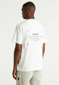 T-shirt bianco a maniche corte in cotone con stampa grafica sul retro e testo grigio. Fessure laterali e vestibilità rilassata. Design semplice.