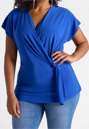 Woman wearing a bright blue wrap-style blouse with short sleeves and light blue jeans, hand resting on hip.