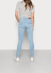 Mujer con jeans ajustados de color azul claro, zapatillas blancas y una camiseta de rayas de manga corta, de pie con la espalda hacia la cámara.