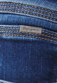 Tela de mezclilla azul con costuras amarillas y una pequeña etiqueta metálica que dice "Pepe Jeans" en el borde de un bolsillo.