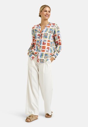 Frau trägt bunt gemustertes Langarmoberteil, weiße weit geschnittene Hose und beige Sandalen mit Schnalle, steht mit den Händen in den Taschen und lächelt.