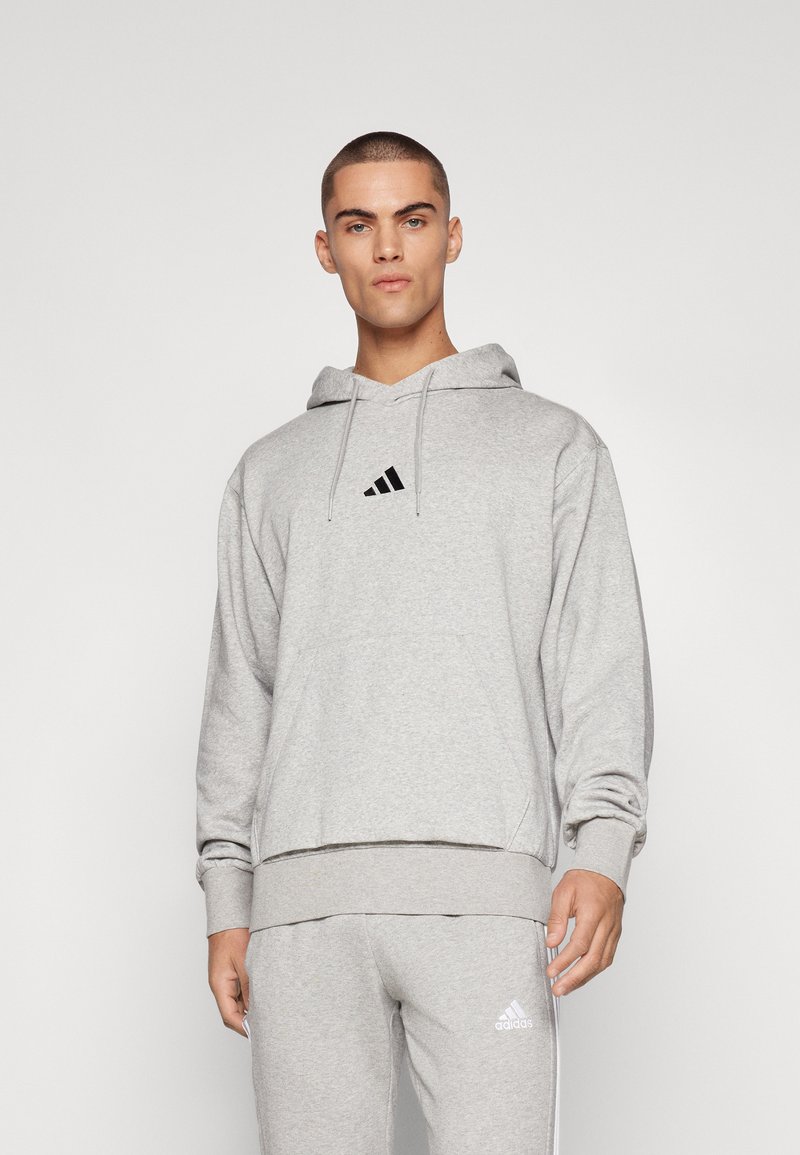 Hoodie cinza claro feito de tecido suave, apresentando um bolso frontal, capuz com cordão e um logo preto da Adidas no peito.