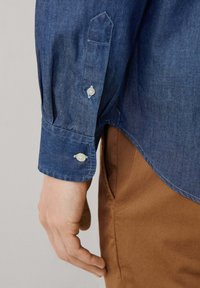 Donkerblauwe spijkerblouse met een smalle manchette met twee witte knopen, ontworpen met een klassieke snit en een soepele textuur. Bruine broek zichtbaar daaronder.