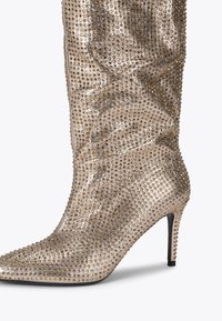 Kniehoge stiletto laars in metallic gouden stof, bedekt met gouden studs. Voorzien van een spitse neus en een strakke, slouchy schacht.