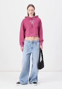 Felpa corta rosa con tasca frontale, abbinata a jeans a gamba larga blu chiaro. Borsa nera indossata sulla spalla, sneakers bianche.