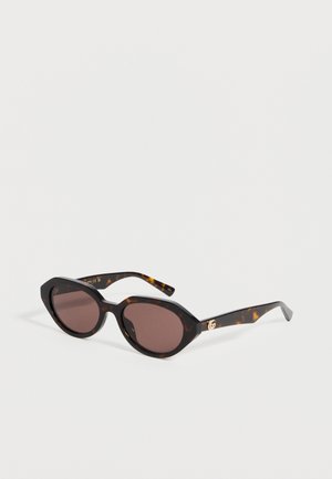 Lunettes de soleil à monture écaille avec verres ovales foncés et détail discret du logo doré sur les branches, présentées sur une surface blanche.