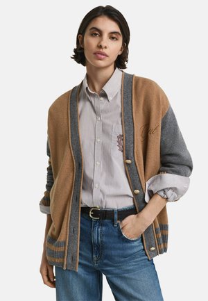 Jonge vrouw in een grijs en beige cardigan over een gestreept overhemd en een blauwe spijkerbroek, met één hand in haar zak.