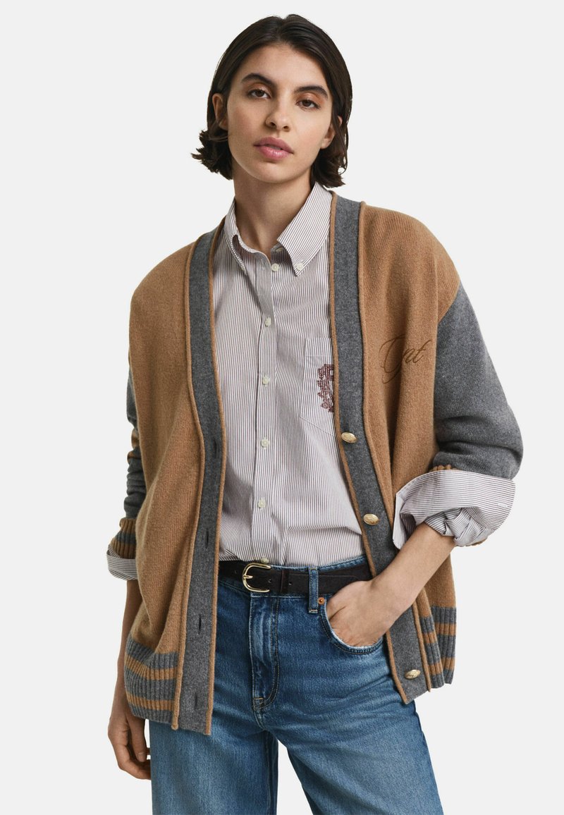 Jeune femme portant un cardigan beige et gris sur une chemise à rayures et un jean bleu, avec une main dans sa poche.