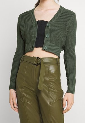 Femme portant un cardigan court en maille vert foncé sur un top court noir et un pantalon taille haute vert olive ceinturé.