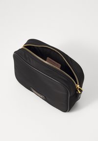 Borsa nera in nylon con zip dorata, forma rettangolare, texture liscia e un'etichetta in pelle con logo sul davanti. Aperta per rivelare il compartimento interno.