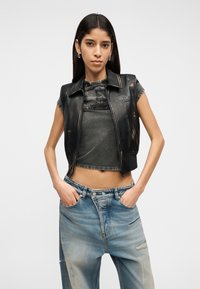 Νεαρή γυναίκα με μακριά σκουρόχρωμα μαλλιά φοράει αμάνικο μαύρο δερμάτινο γιλέκο, γκρι κοντό τοπ και φαρδιά ξεθωριασμένα μπλε τζιν με τα χέρια στις τσέπες.