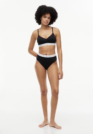 Γυναίκα με σγουρά μαλλιά που φοράει μαύρο bralette Tommy Hilfiger και εσώρουχα με ψηλή μέση, στέκεται ξυπόλητη σε λευκό φόντο.