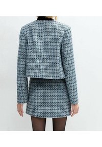 Kvinna klädd i en blå och svart rutmönstrad tweedjacka och matchande kjol, stående med armarna avslappnat längs sidorna, med ryggen mot betraktaren.