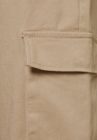 Un tissu beige à la texture lisse présente une grande poche avant et des coutures visibles. Le design met l'accent sur l'utilité et la simplicité.