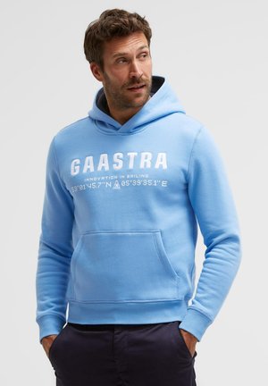 Man wearing light blue Gaastra hoodie with white text and black pants, hands in pockets, looking to the side against a plain background.