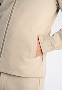Beige zip-jacka i mjukt tyg, med en ficka på sidan och slät textur, med rak fåll och sydda detaljer.