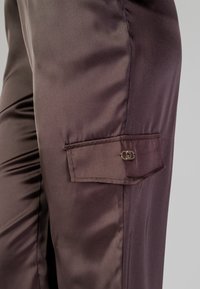 Pantaloni in satin scuro con una texture liscia, dotati di una tasca laterale con chiusura a bottone e una lucentezza sottile.