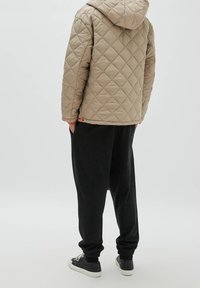 Veste matelassée beige avec capuche, associée à un pantalon de survêtement noir. Détails remarquables : poignets élastiques et accents orange contrastants.