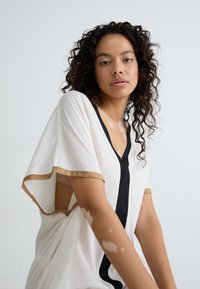 Loose-fitting cream blouse featuring a black vertical line and gold trim on sleeves, with a deep V-neckline and open side design.