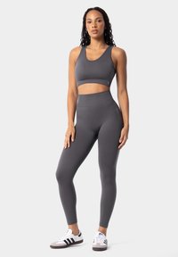 Šedý športový crop top a legíny s vysokým pásom vyrobené z elastického, hladkého materiálu. Model má na sebe šedé a biele tenisky s čiernymi pruhmi.