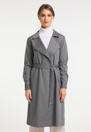 Frau trägt einen grauen Trenchcoat mit Gürtel über einem weißen Rollkragenpullover, stehend vor einem einfarbigen hellen Hintergrund.
