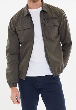 Homme portant une veste zippée olive sur un t-shirt blanc et un pantalon bleu foncé, debout avec la main gauche détendue sur le côté.
