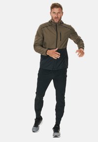 Herrenjacke in Olivgrün und Schwarz, mit einem Reißverschluss vorne, Kapuze und kontrastierenden Einsätzen. Getragen mit schwarzen Sporthosen und Schuhen.