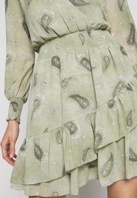 Grünes Paisley-Kleid aus leichtem Stoff, mit gesmoktem Taillenbereich, langen Ärmeln und schichtweise Rüschen am Saum.